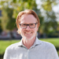 Smiling man with glasses and red hair standing outside in a park-like setting on a sunny day.